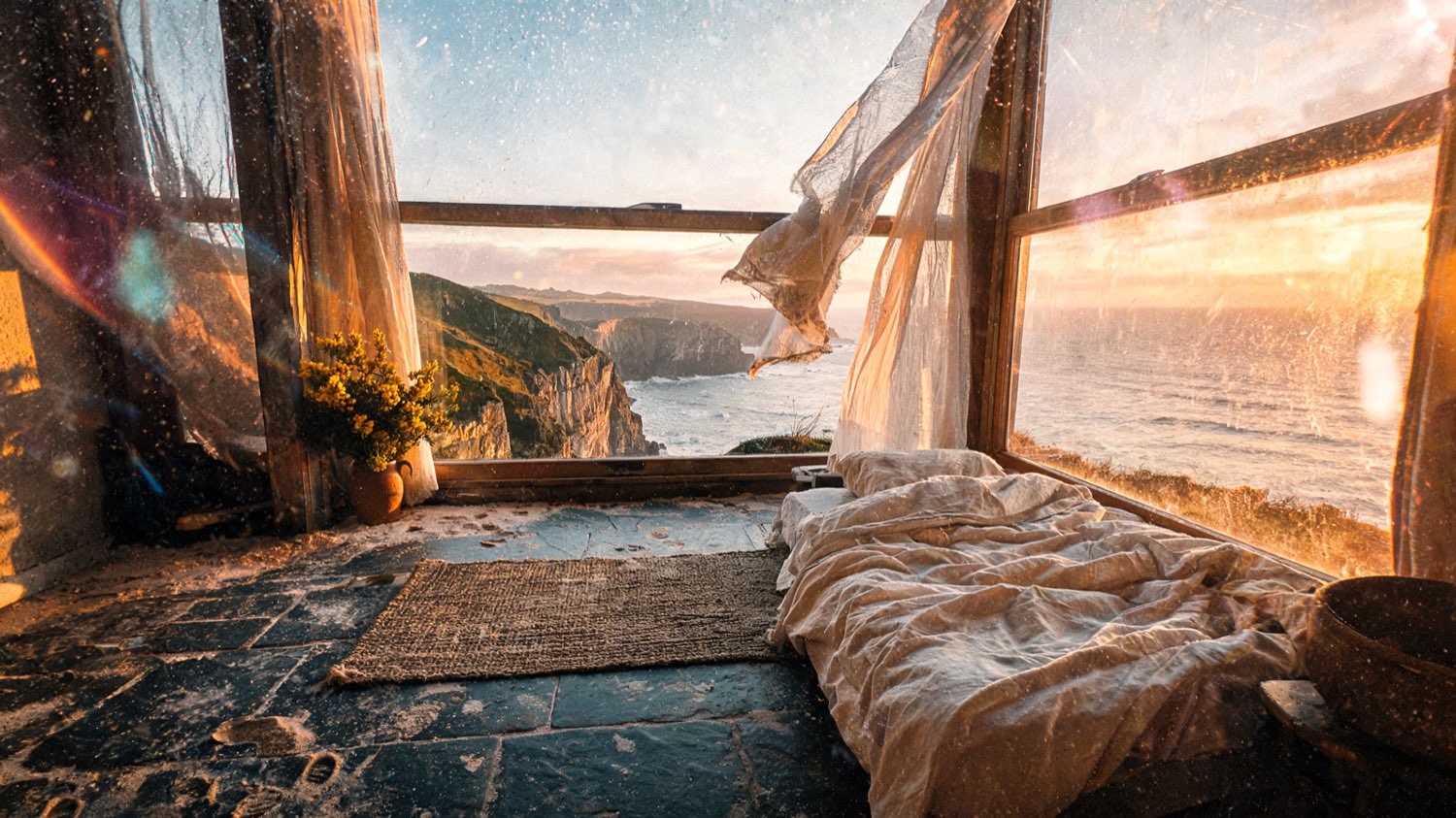 Suite interior with a low linen bed, woven sea-grass rug and tall windows open to the Cornwall sea and headland